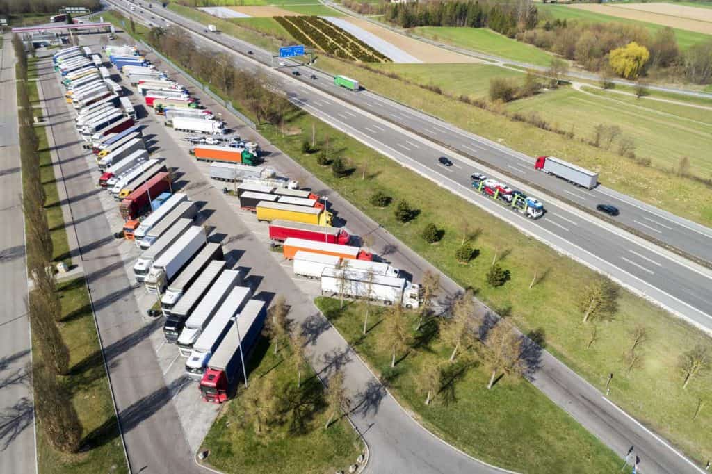 Aerial view of a multiple lane highway and a large truck stop with long rows of parked semi trucks.