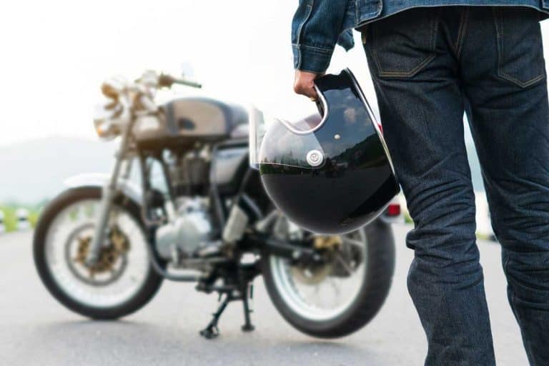 Motorcycle rider in jeans holding a helmet with his motorcycle blurred on the background, How Many Years Does a Motorcycle Helmet Last?