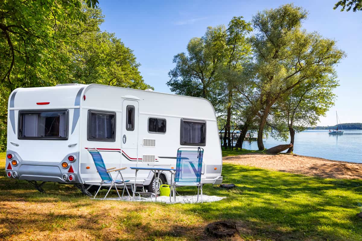 A travel trailer with chairs set up for camping and parked near a lake with a scenic view, Do RV Batteries Charge When Plugged Into Shore Power?