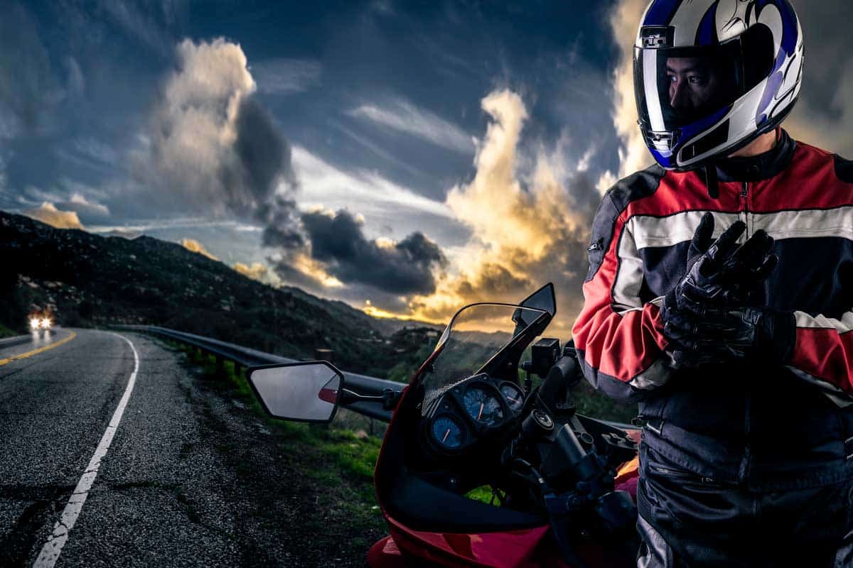 Male motorcyclist wearing protective leather racing suit with a red bike or motorcycle on an open road, What's The Average Cost For Motorcycle Gear?