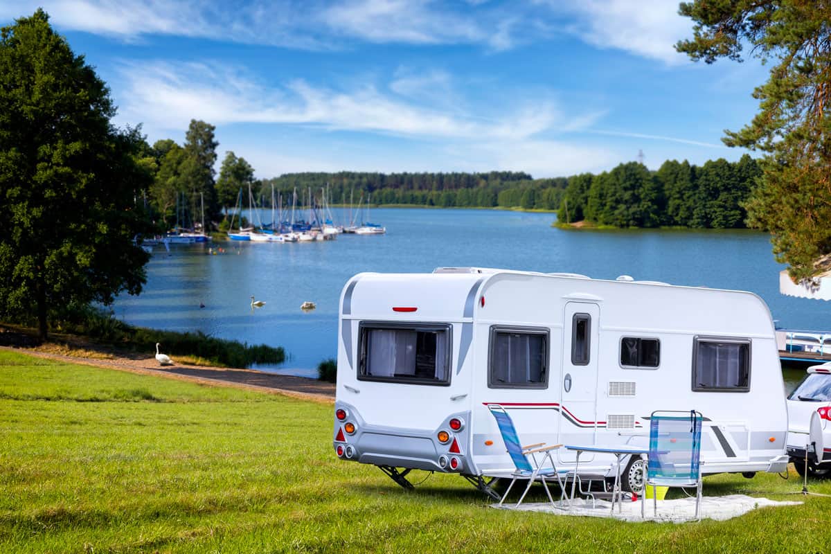 A travel trailer parked on the side of a lake providing a panoramic view of the lake and forest trees, 8 Best Hybrid Travel Trailers For 2021