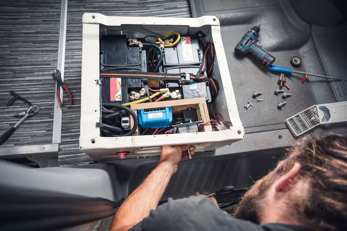 Bearded man working on a DIY camper van. He is working on the base of a front seat to install additional batteries, How Long Will An RV Battery Last (Inc. When Dry Camping)?