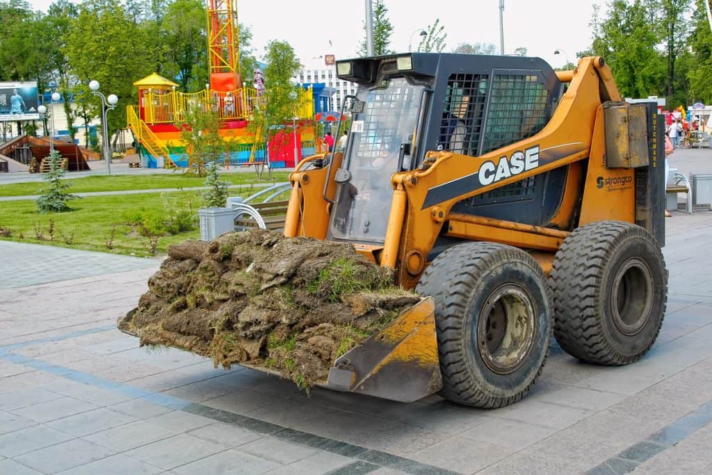 A yellow skid loader hauling grass in a small park