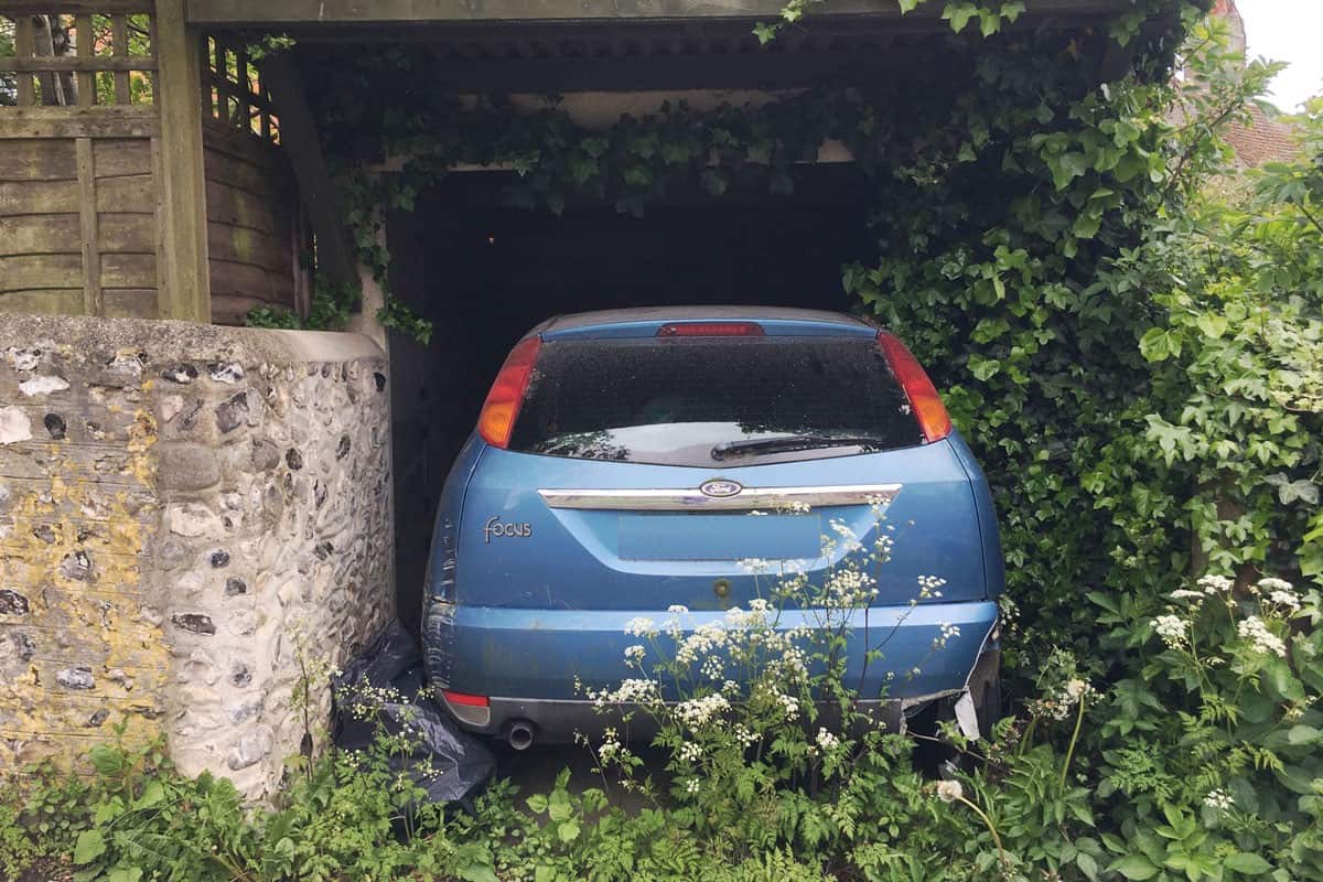 blue car parked in outdoor garage with overgrown weeds indicating that it is unused for a long time during coronavirus pandemic lockdown