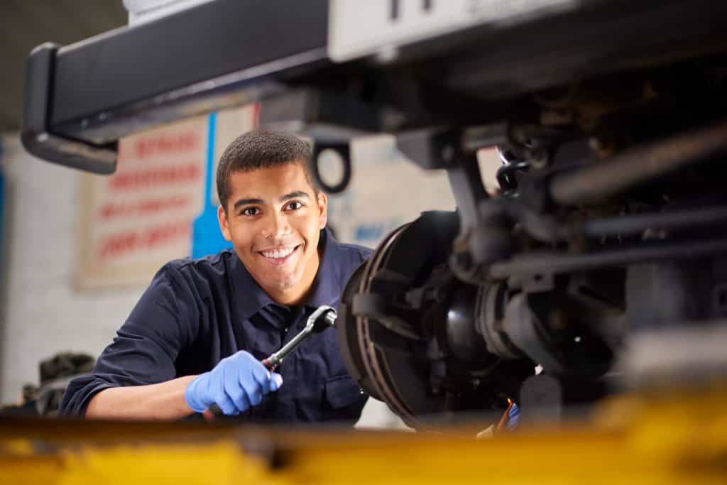A young mechanic is smiling to camera as he works on a car in a garage repair shop. He is wearing blue overalls. He is fixing the brake discs on the front driver's side of the vehicle.