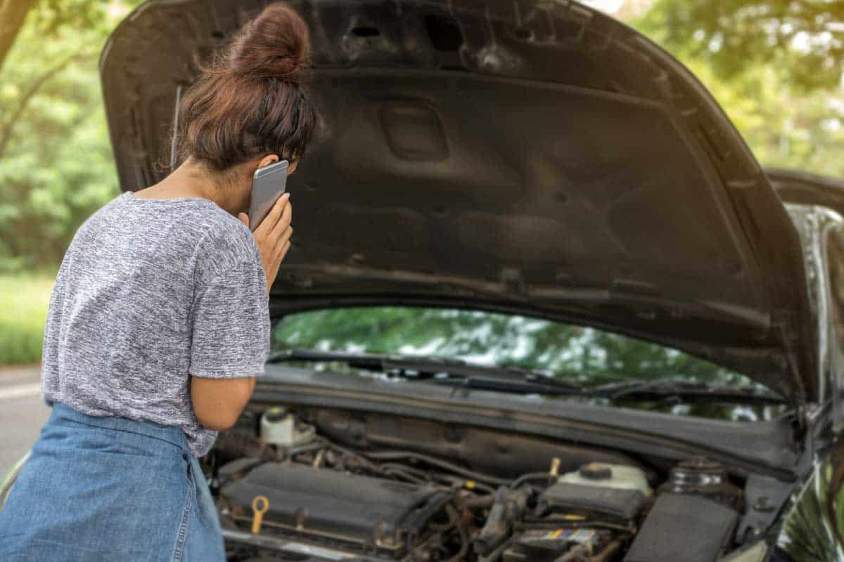 Woman using mobile phone while looking at broken down car on street, Engine Stopped While Driving - Here's What Could Be Wrong