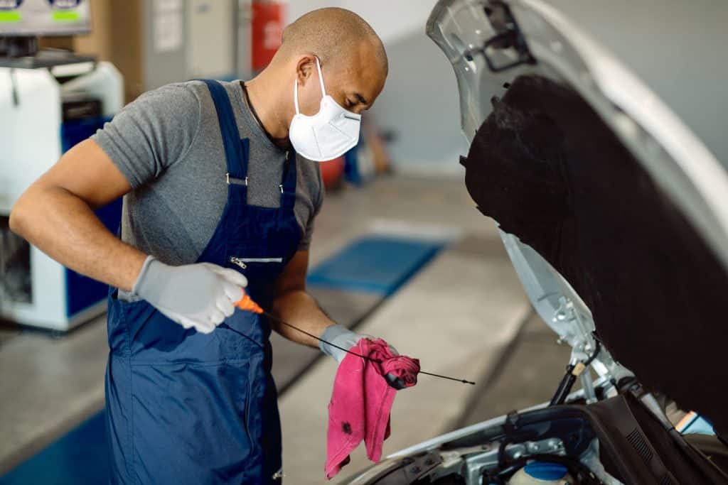  repairman wearing protective face mask and examining car oil in a workshop.