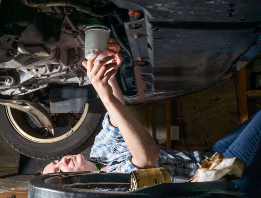 A young woman lies on garage floor replacing the oil filter on her car after performing routine oil change