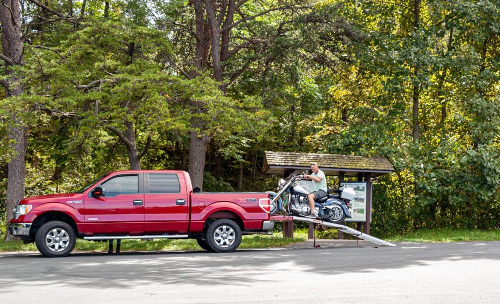 An elderly motorist is unloading his motorbike from the trunk of a red Ford F150 XTR pickup by slowly backing up on a metal makeshift ramp at Shenandoah Park.