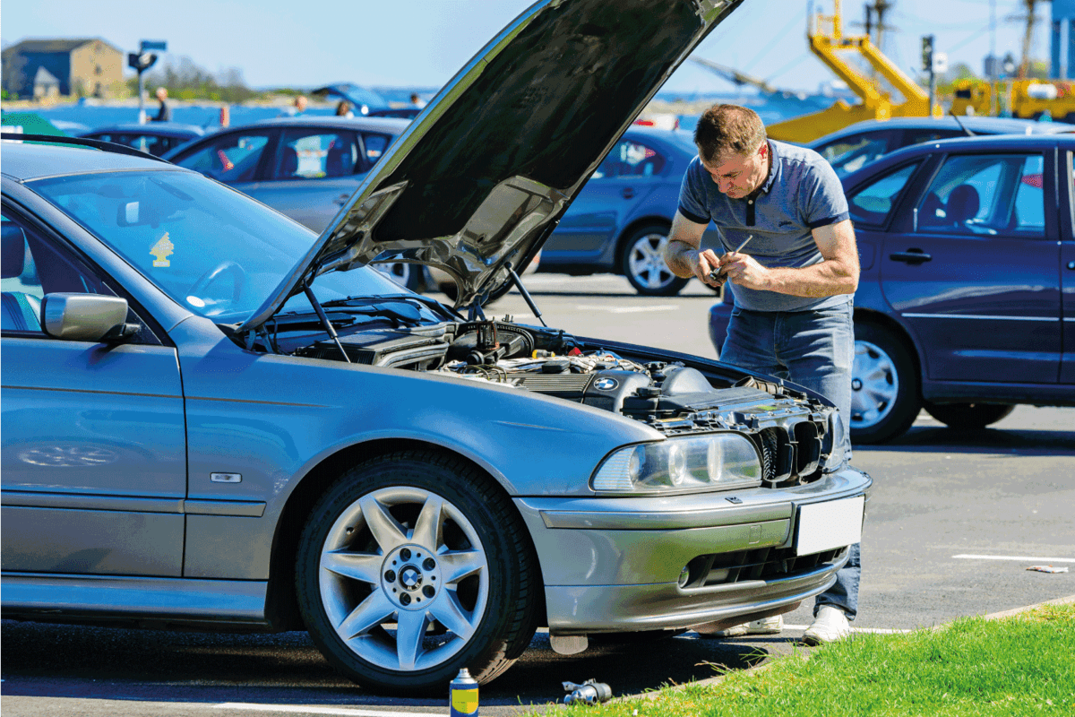 Male person working under the hood of a 2005 BMW 525 touring on a parking lot in the harbor. Hood is open and person is using tools on the motor.