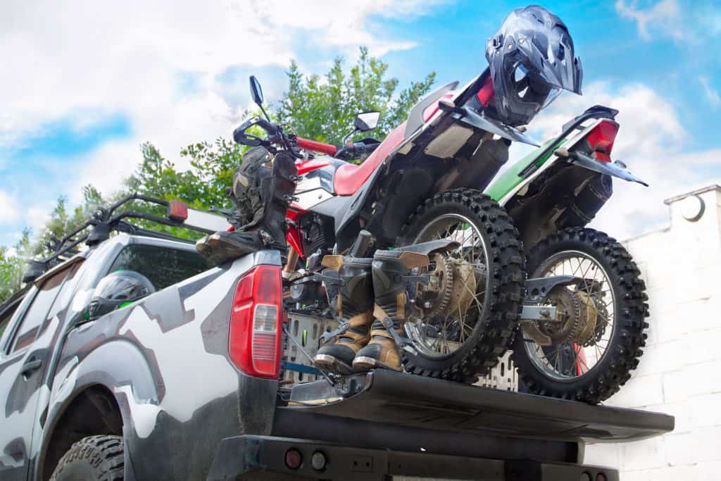 Two dirt bike motorcycles on the back of the camo truck with safety equipment, such as motorcycle boots and helmets, Will A Motorcycle Fit In A Truck Bed?