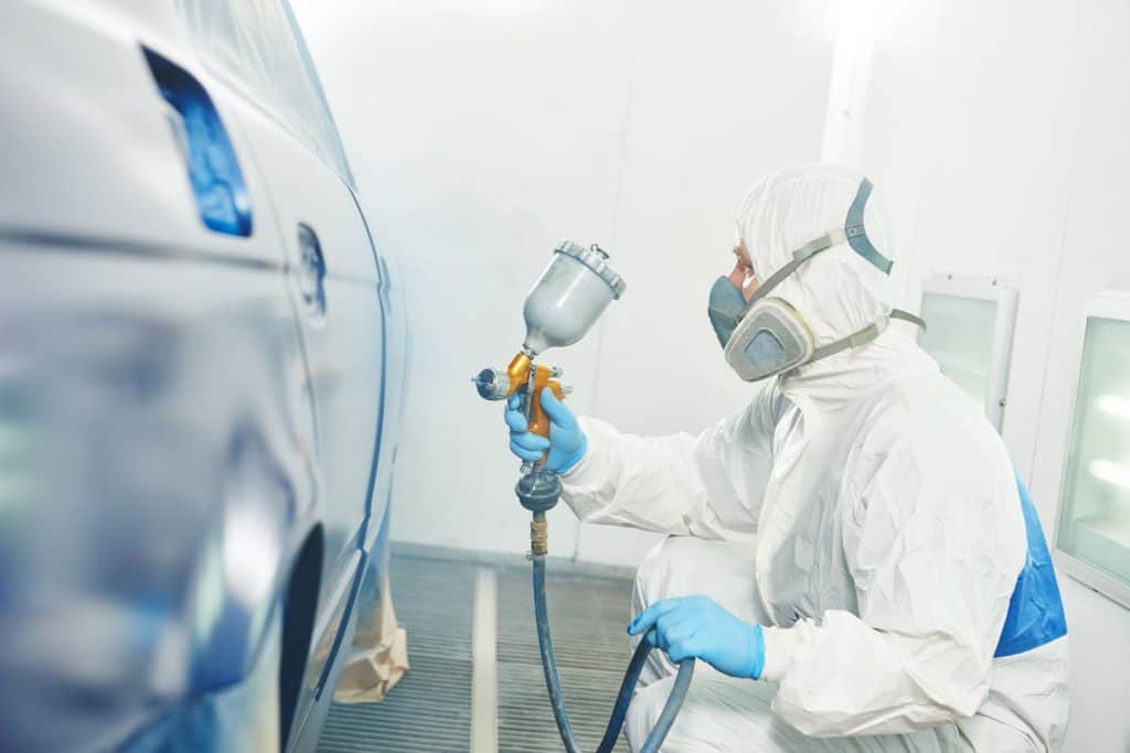 automobile repairman painter in protective workwear and respirator painting car body in paint chamber