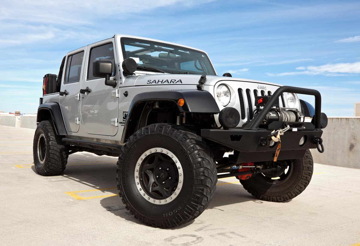 A parked silver 2008 Jeep Wrangler, this particular Jeep has a custom shock kit and winch and is known as the Sahara edition.