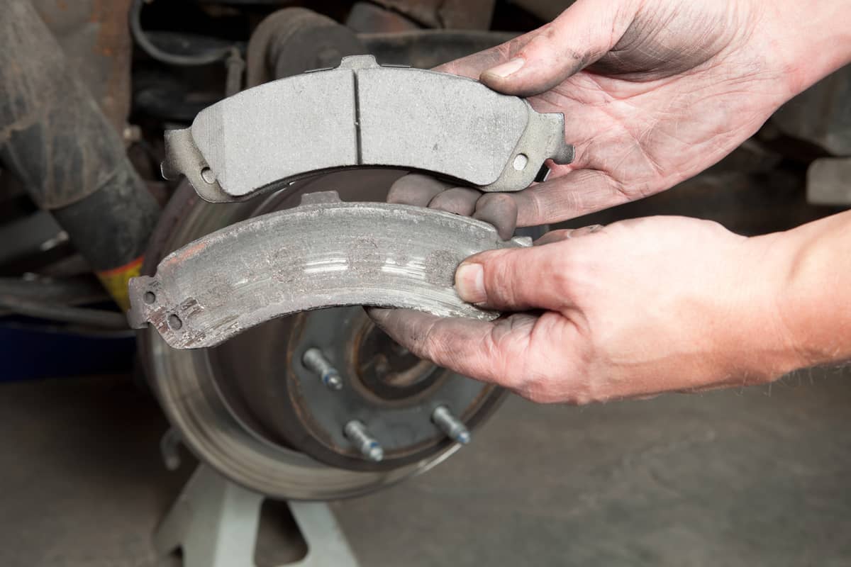 A mechanic's hands are displaying a worn to the metal brake pad (lower) and a new pad (upper). 