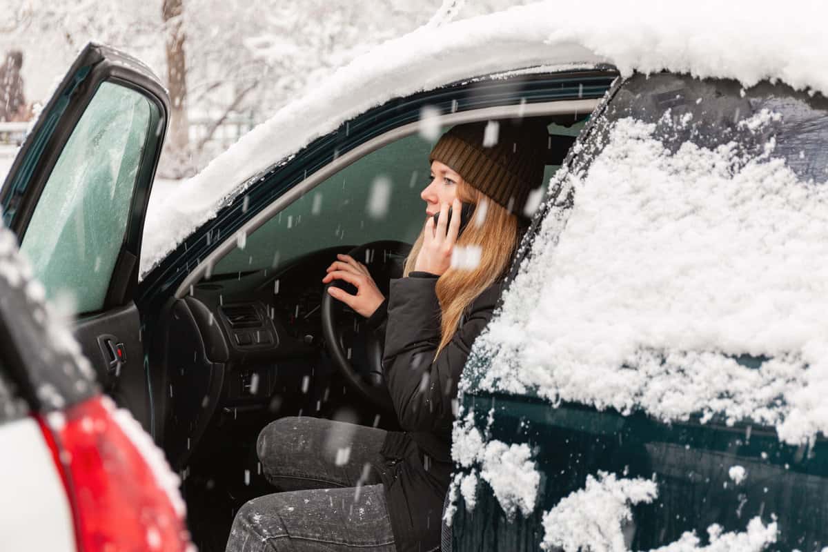Young woman calling for help or assistance inside snow covered car. Engine start in frost. Breakdown services in the winter.