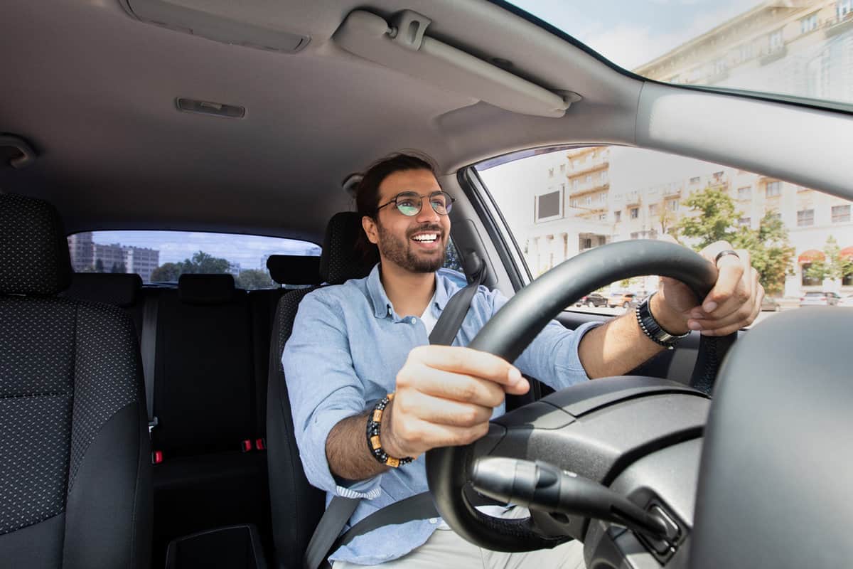 Joyful indian man driving car, shot from dashboard, going on trip during summer vacation. Happy middle-eastern guy in casual outfit and glasses driving his brand new nice car