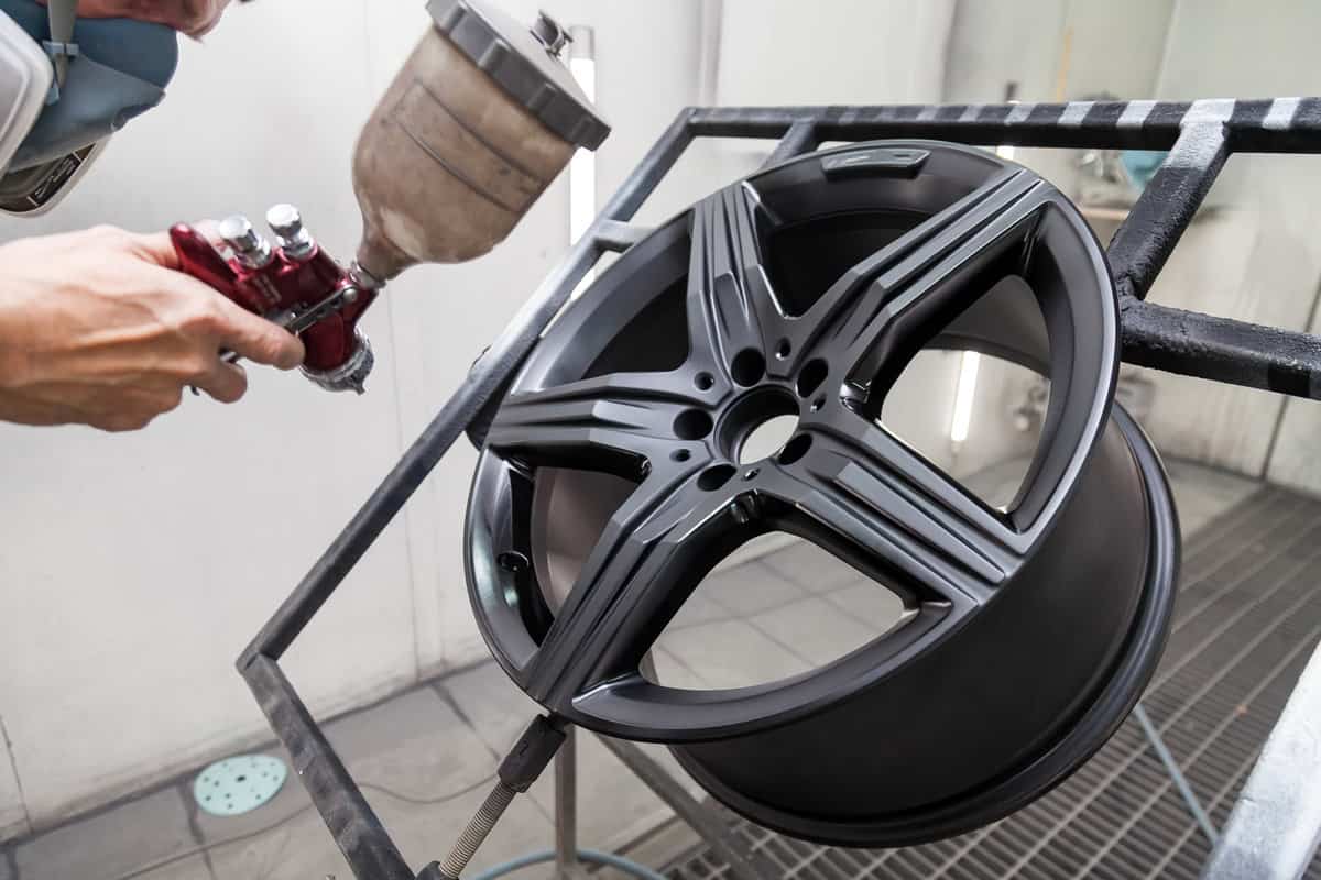 Painting the element body of the car - the aluminum alloy wheel with the help of aerograf in black color by the hand of painter repairman in the industrial professional garage. Auto service industry.