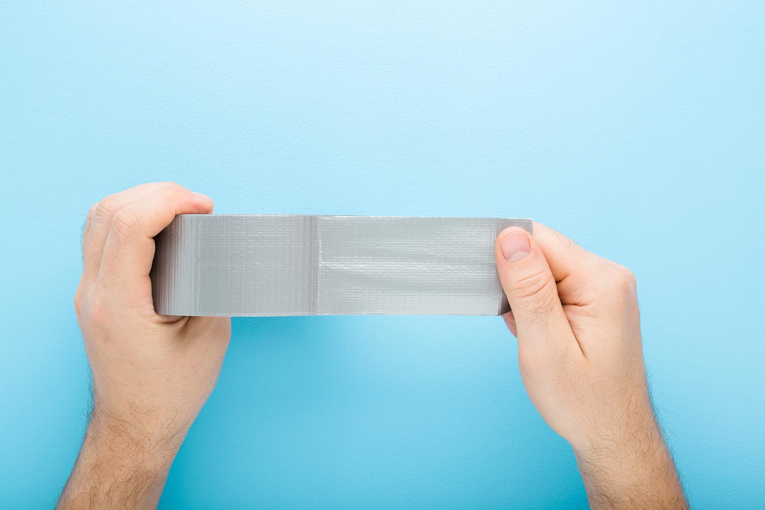 Young adult man hands stretching gray adhesive tape on light blue table