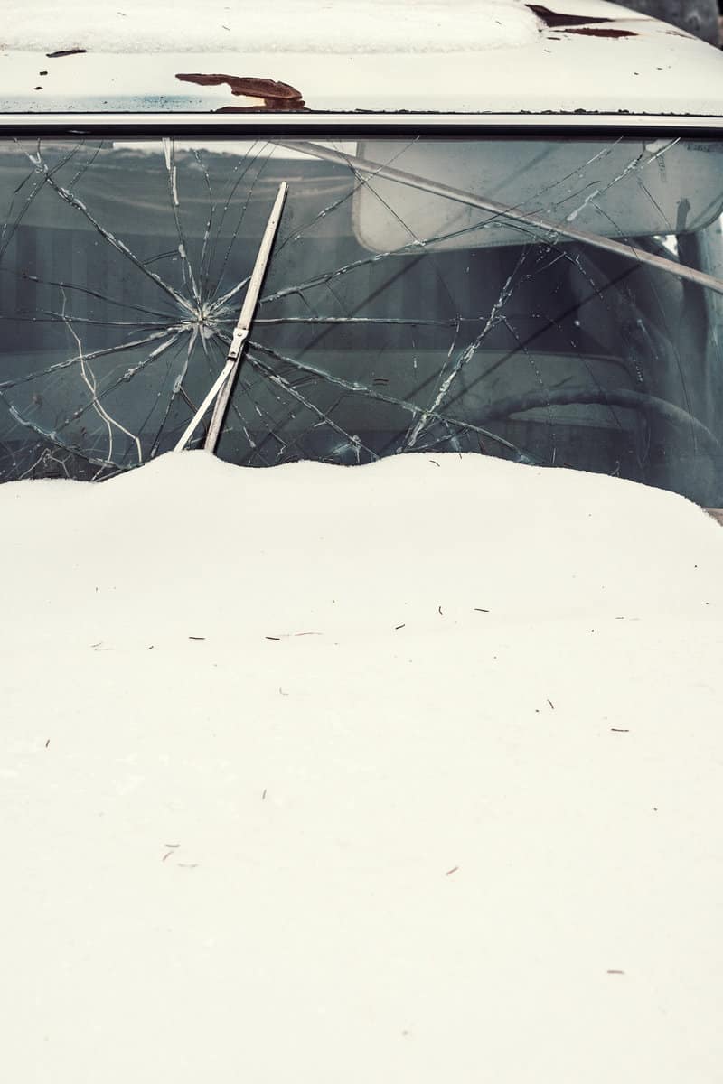 A 1960's sedan covered in snow with a cracked windshield.
