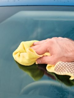 Close up mans hand polishing or cleaning a front windshield of a blue automobile with a yellow microfiber rag . Manual car washing - How To Remove State Farm Beacon From Windshield