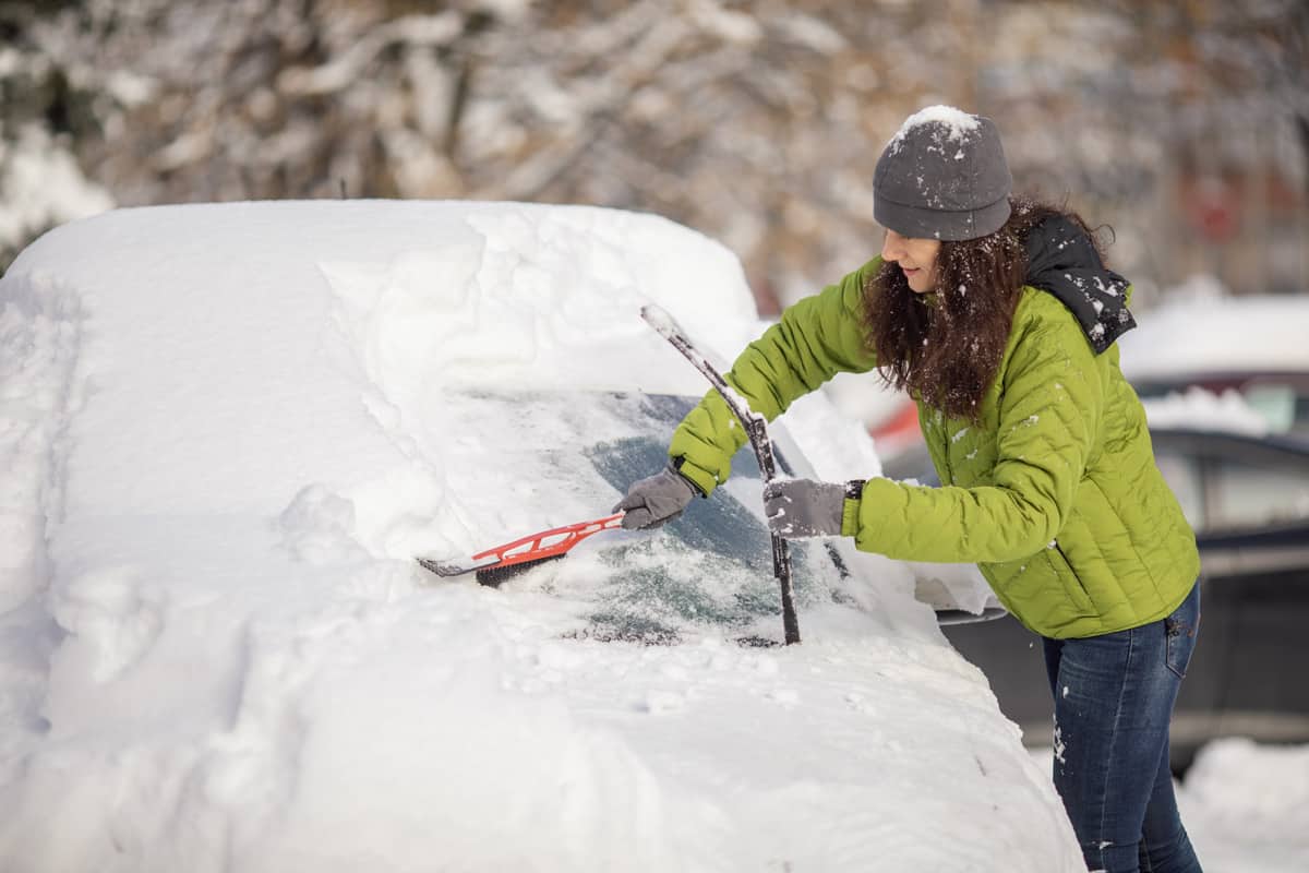 Mature aged woman cleaning her car from snow on parking. She wears warm clothes and using brush for snow