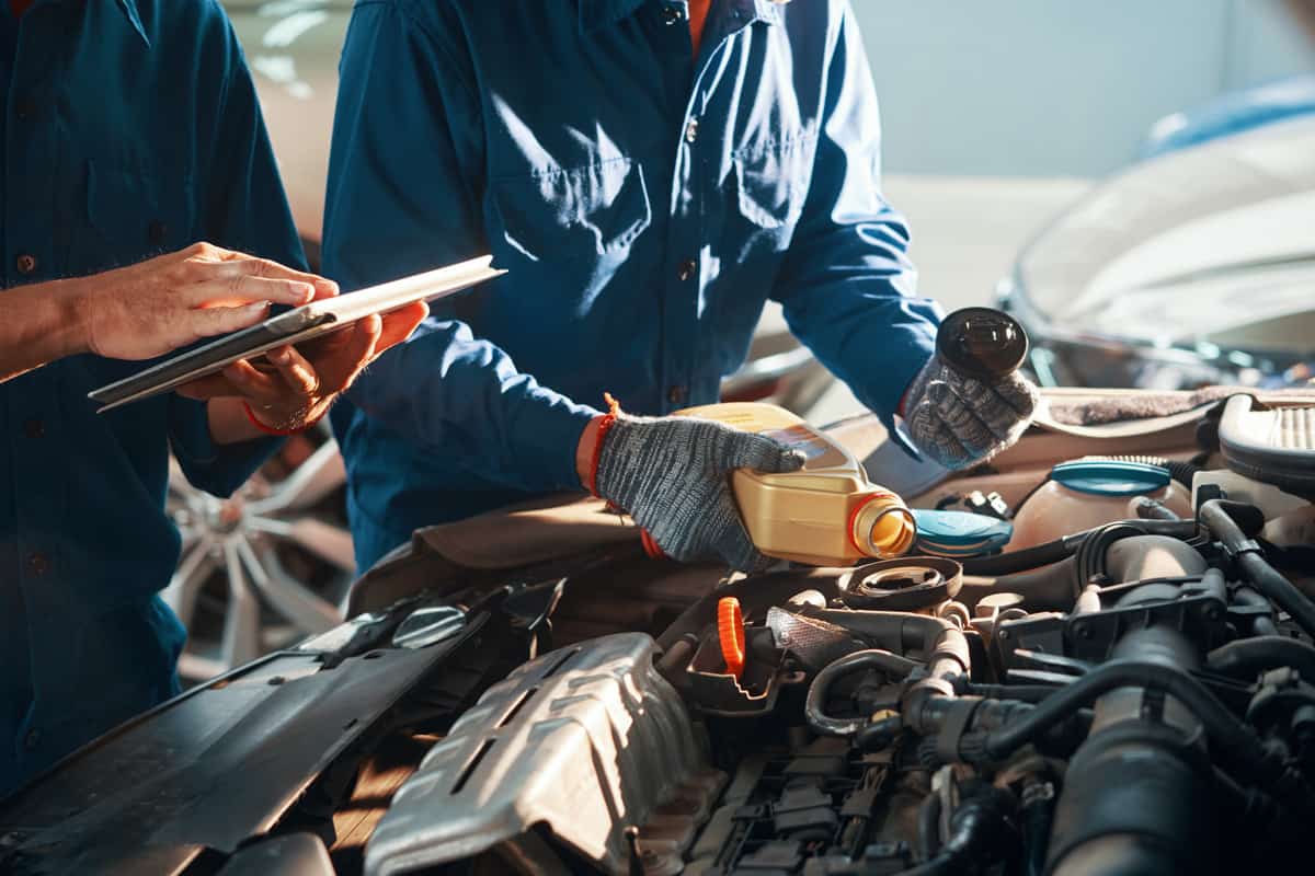 Mechanic pouring oil in car engine when his colleague using tablet computer