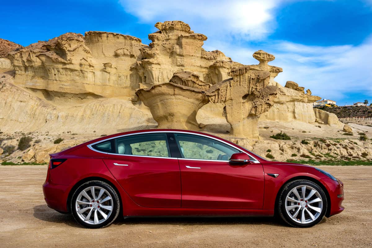 a red Tesla Model 3 electric car with rock formation on the mediterranean coast and blue sky