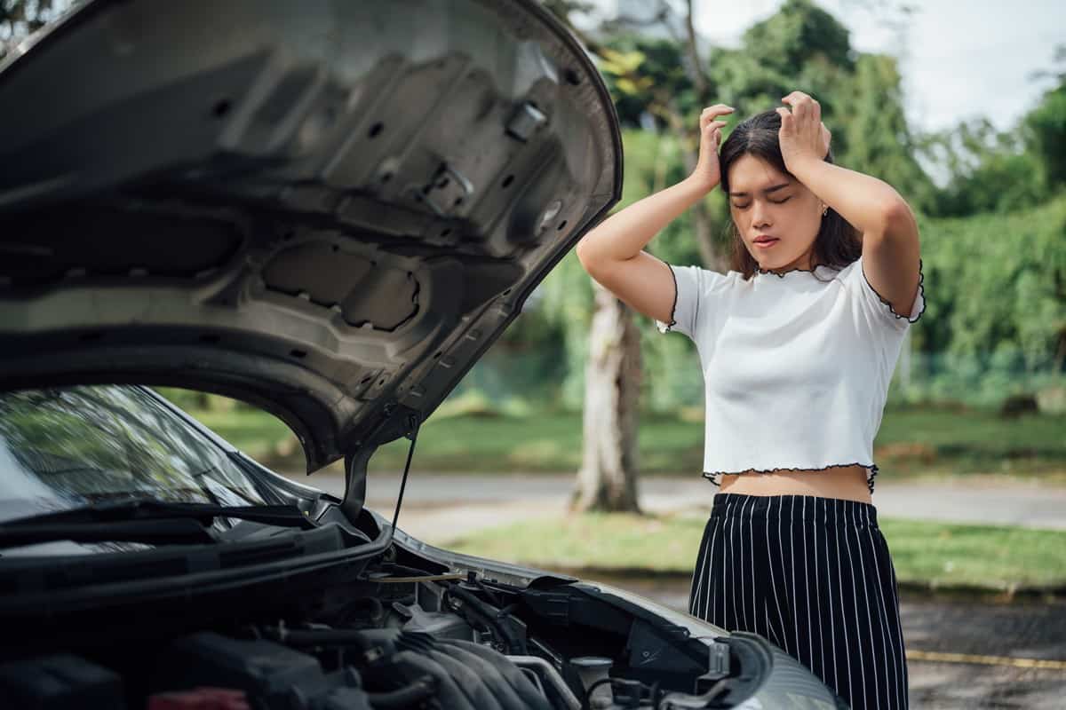 Confused beautiful Asian female driver in casual wear looking at broken down car engine under hood on the street during road trip, looking puzzled and helpless.