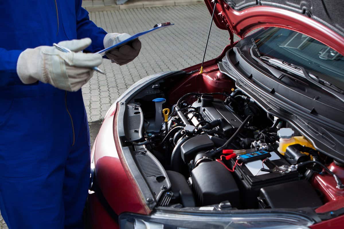 Mechanic Holding Clipboard In Front Of Open Car Engine — Photo