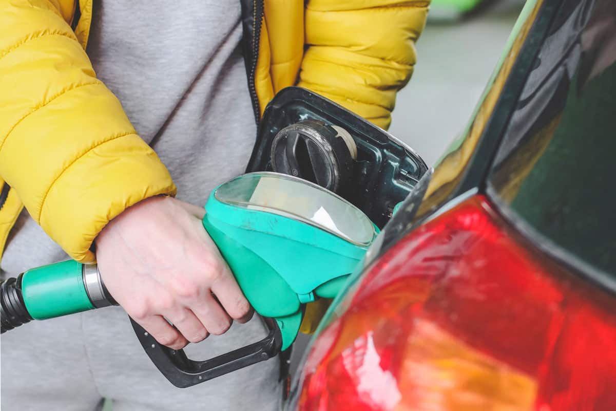 The hands of a young caucasian man in a yellow jacket hold a refueling gun in the tank on a green car