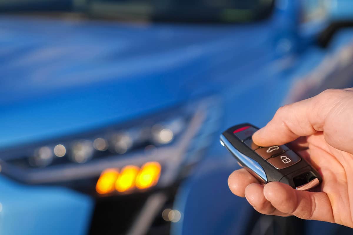 Man uses thumb to press black button on remote control key removing alarm signaling from bright blue car and headlights flash