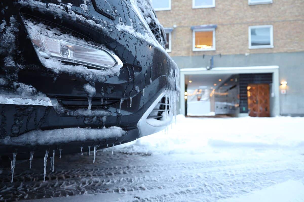Front of the unrecognizable car covered with snow and ice. Somewhere in the city of Halmstad. There is an unrecognizable random building at the background, Surface of the road is covered with snow.