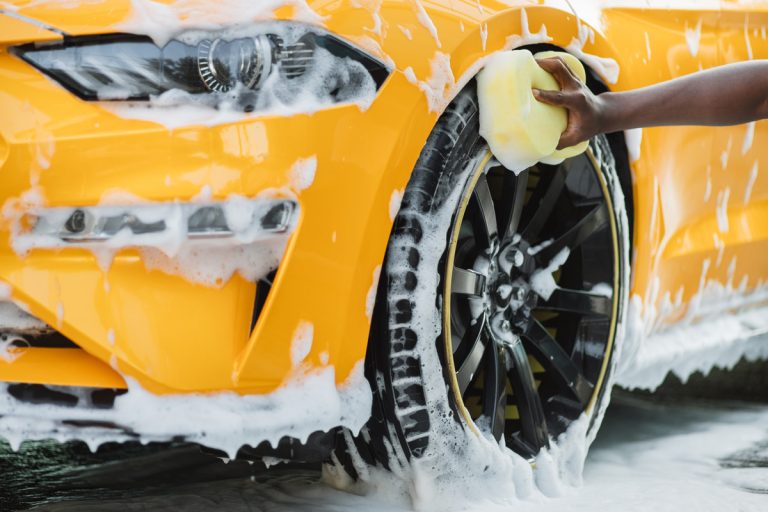 Hands of African man holding yellow sponge, washing car wheel with foam. Cleaning of modern rims of luxury yellow car at self car wash service outdoors., Can You Wash A Car With Laundry Detergent?