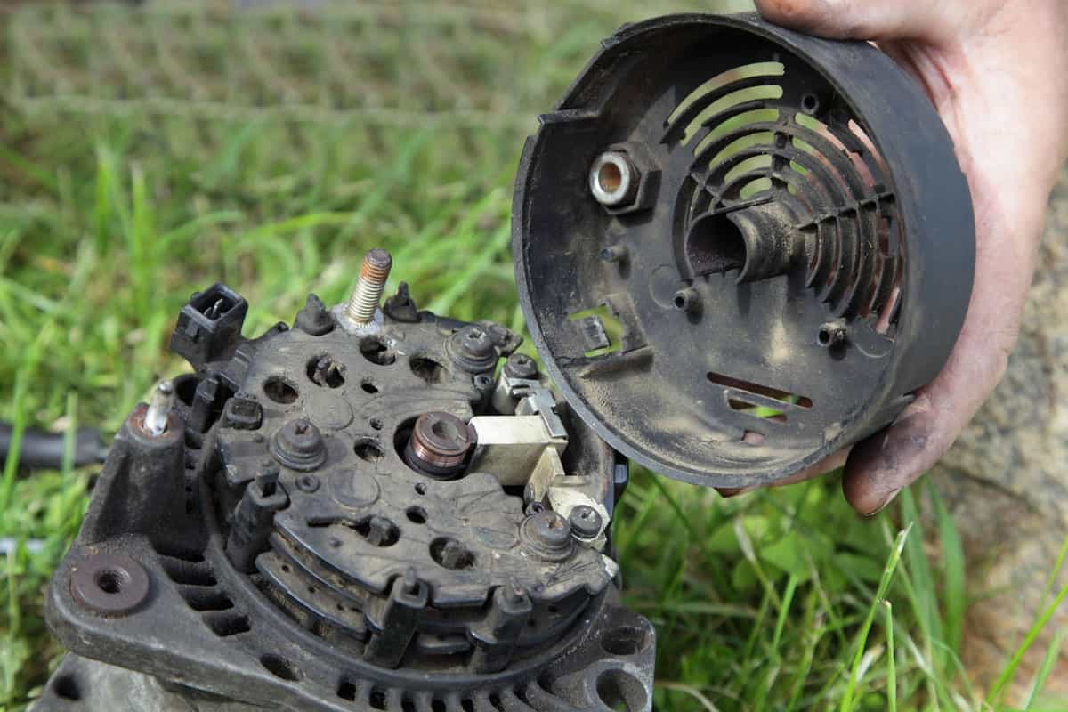 Used car 12 V alternator close up with removed back cover in hand on green grass background, outdoor vehicle electric equipment repair 