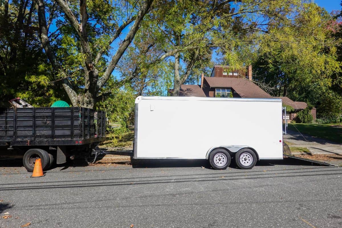 A landscaping truck with a long white enclosed trailer trailer seen on a shady residential asphalt street