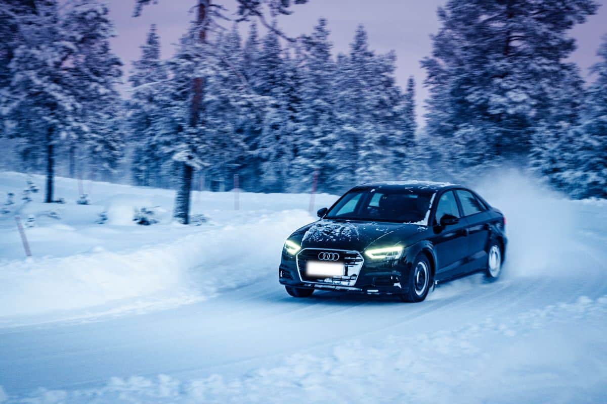 Audi A3 Sedan drives on a snowy road near winter forest at dusk beyond the Arctic Circle. Extreme winter driving conditions. Front LED headlights are on.