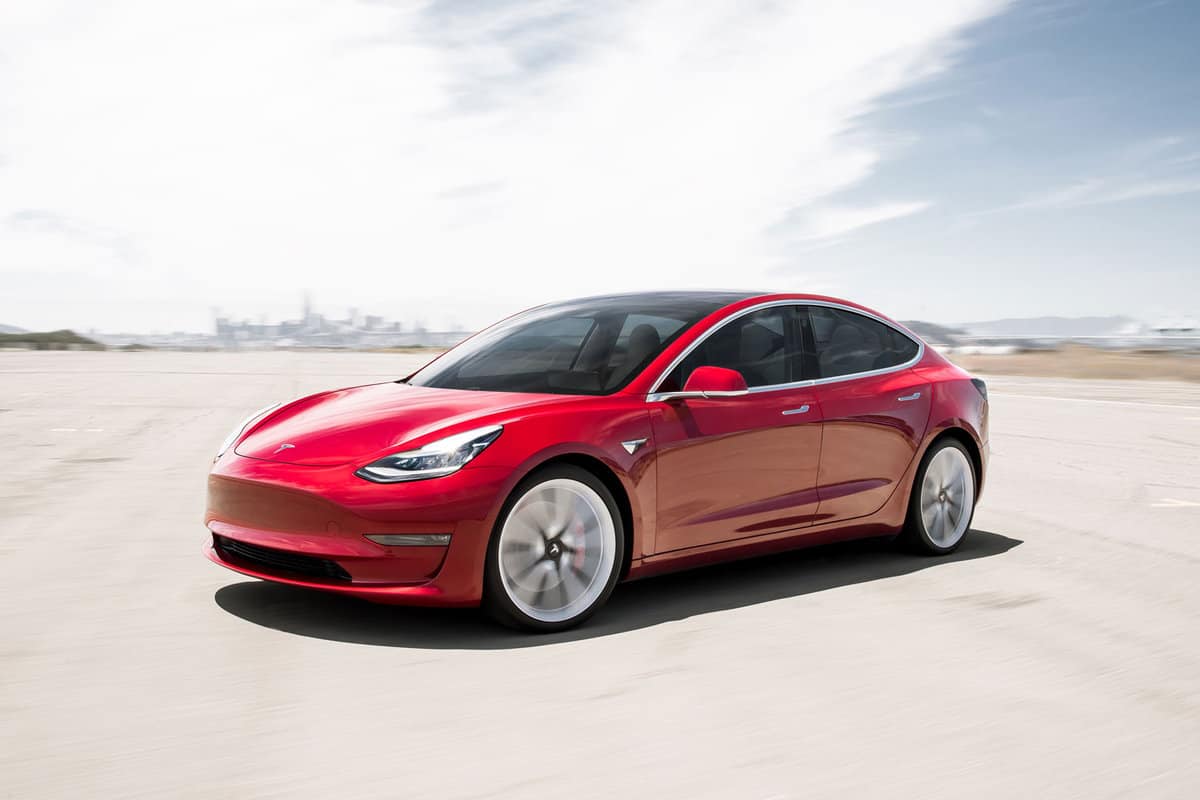 Photograph of a red Tesla model 3 in front of a distant city.