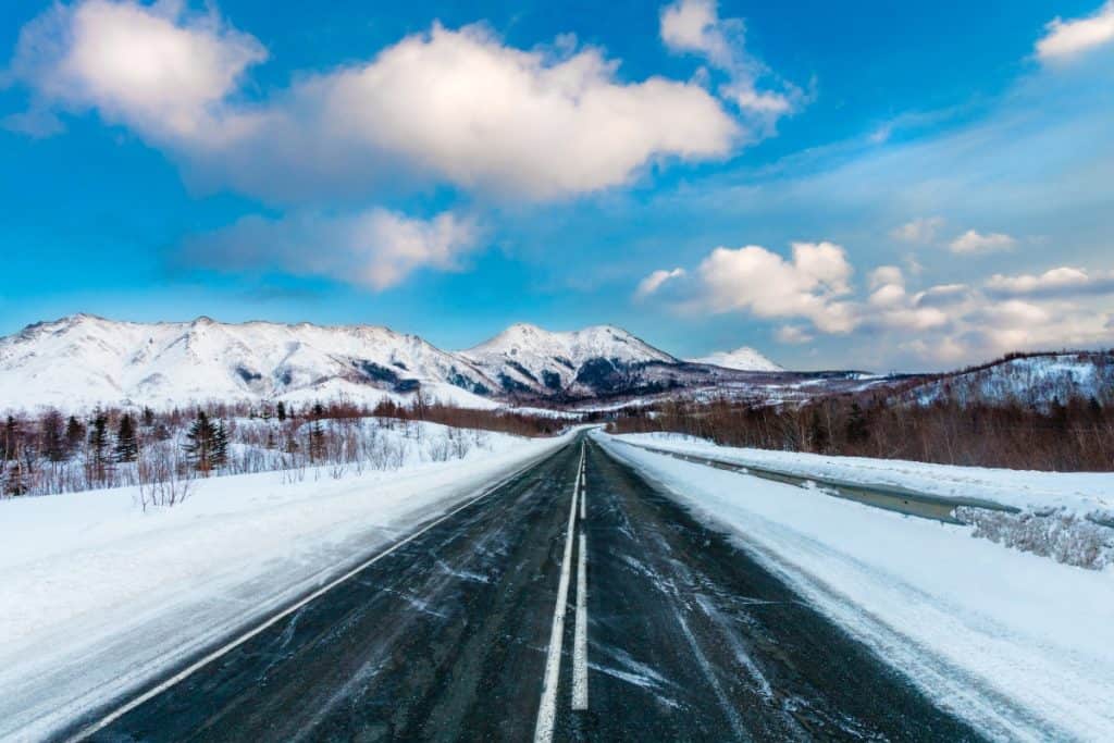 Empty snow covered asphalt dark road with white road marking along mountains and hills and blue sky with clouds. Russia. Island Sakhalin

