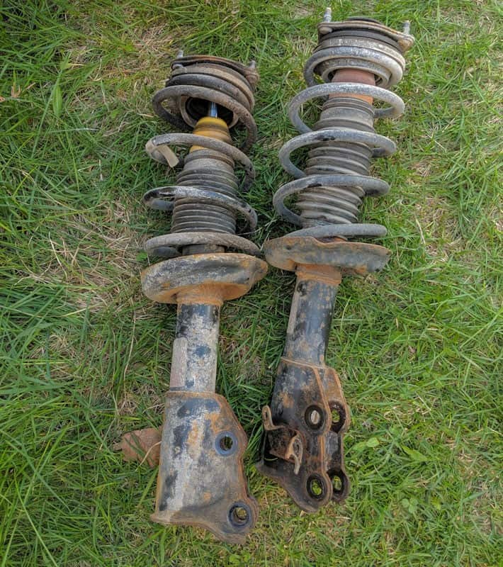 A pair of old rusty coilover struts laying on green grass before disposal.
