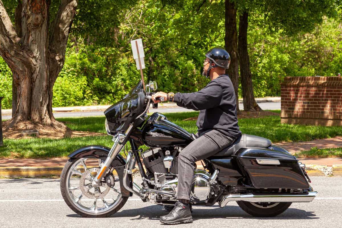 An African American man with goatee beard wearing jeans, jacket, boots and helmet is idling on the road in the city. He is riding a Harley Davidson motorbike.
