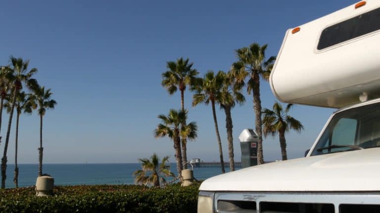 An image of a motorhome with a beautiful background of california, palm trees, and the ocean.