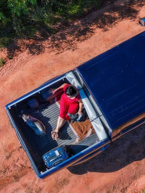 a woman sitting in the back of a pickup truck with her dogs and suitcase, all on a typical red sand road